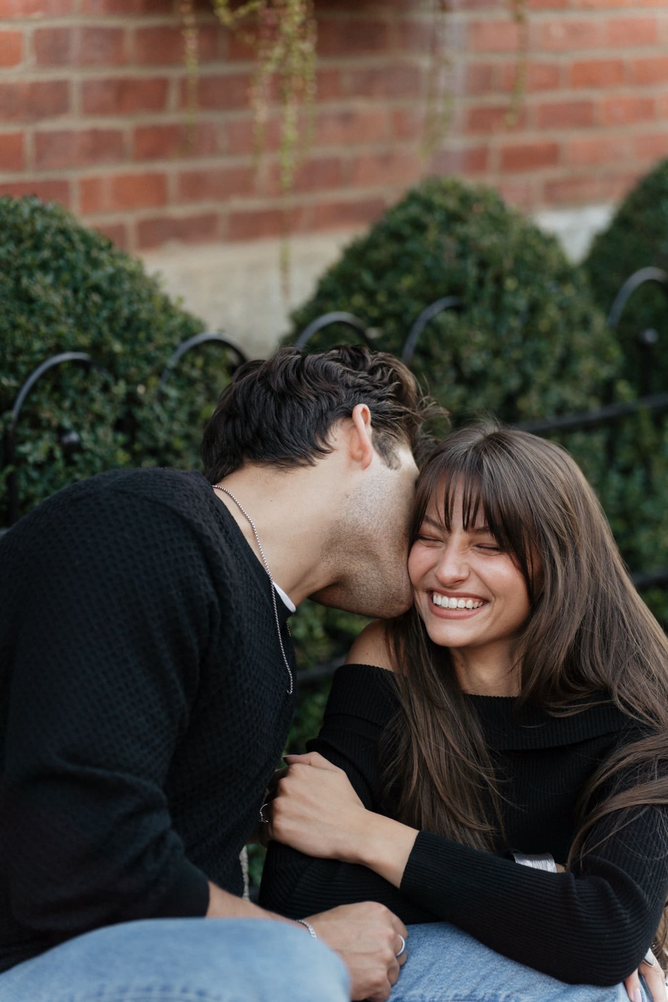 Man kissing his laughing fiancée's cheek beside boxwood hedges in German Village, Columbus, Ohio